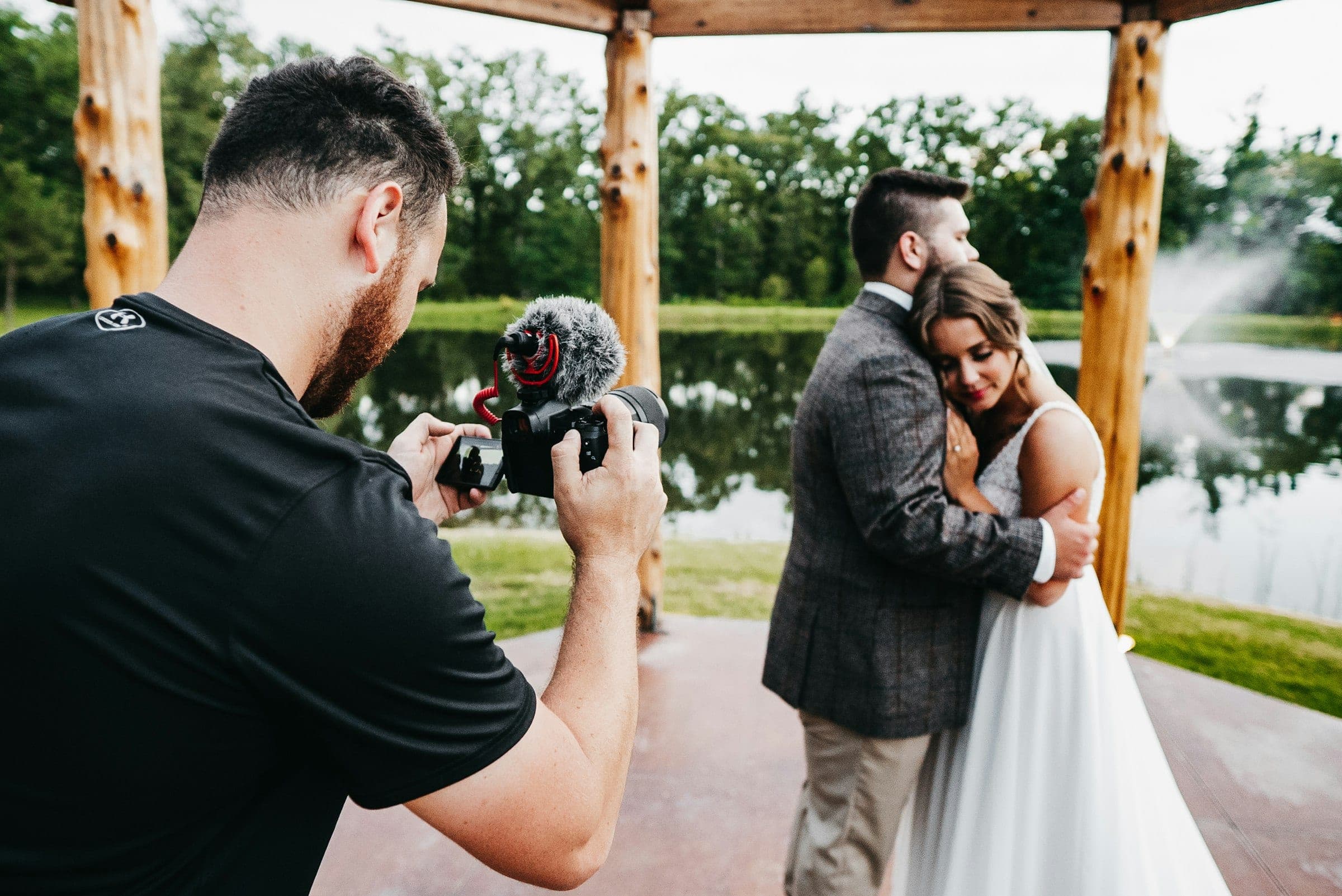 A wedding photographer filming a bride and groom embracing under a timber gazebo by a lake